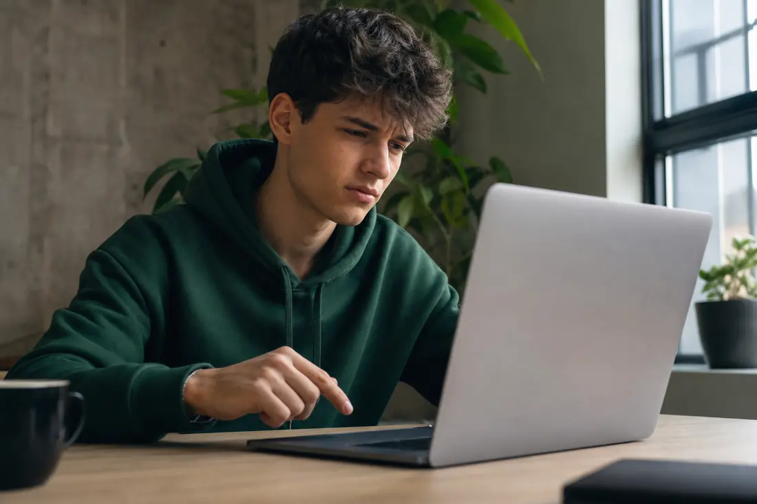Jeune homme en sweat à capuche vert assis à un bureau en bois devant un ordinateur portable, l'index suspendu au-dessus du pavé tactile, l'air méfiant
