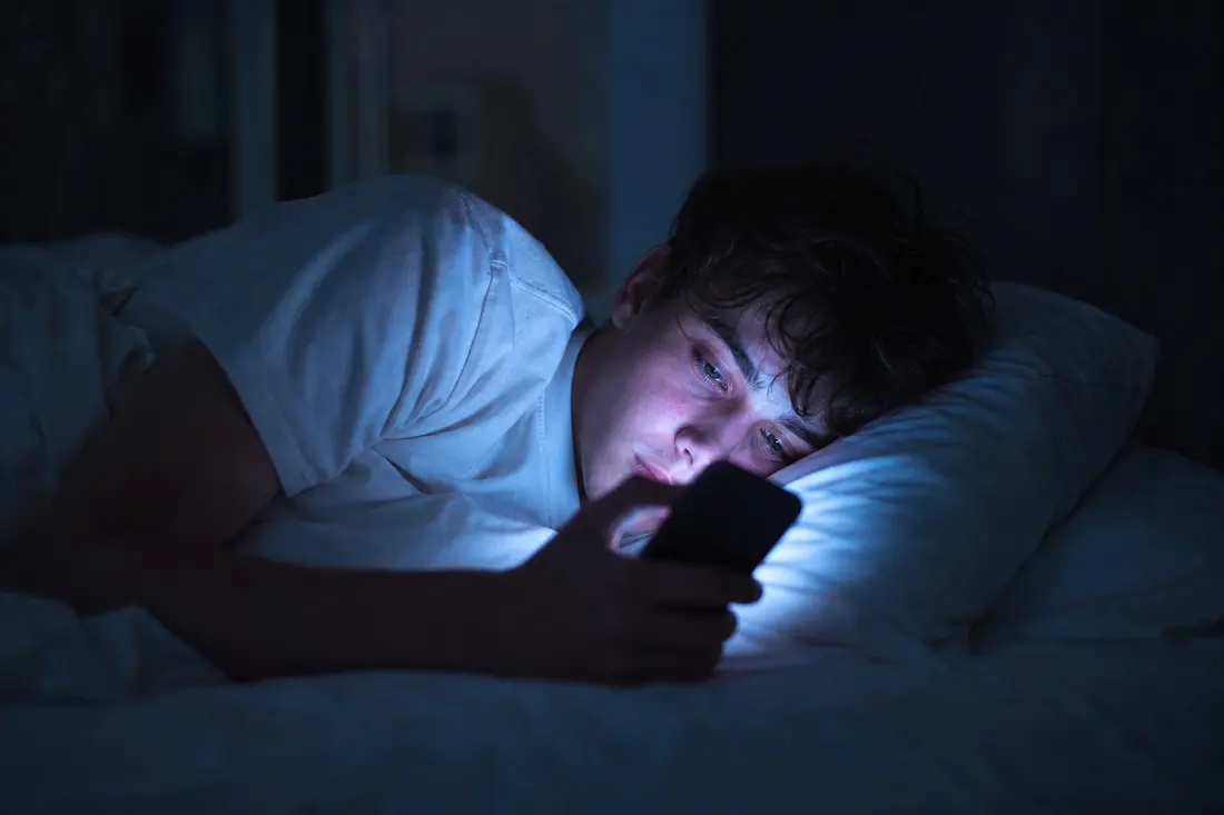 Jeune homme allongé dans son lit dans le noir, la tête sur l'oreiller, le visage éclairé en bleu par l'écran du téléphone qu'il tient devant lui