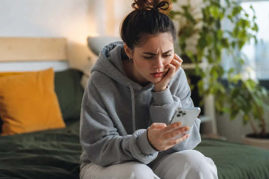 Jeune femme en sweat gris assise sur son lit, la joue posée sur la main, fronçant les sourcils devant l'écran de son téléphone