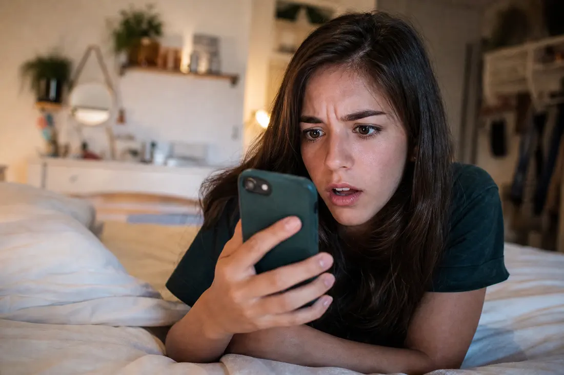 Jeune femme allongée sur son lit dans une chambre tamisée, les sourcils froncés, fixant l'écran de son téléphone d'un air stupéfait