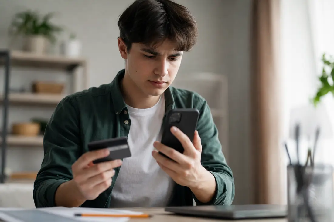 Jeune homme en chemise verte assis à son bureau, carte bancaire dans une main et smartphone dans l'autre, l'air contrarié devant son ordinateur portable