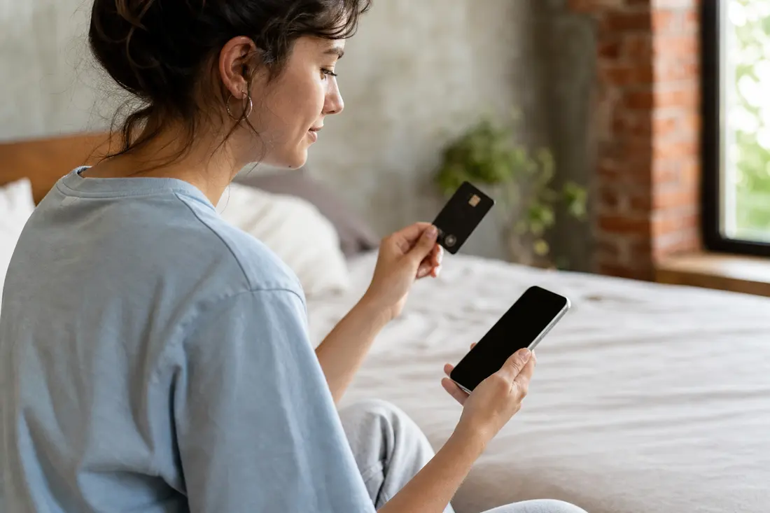 Jeune femme assise sur son lit, carte bancaire dans une main et smartphone dans l'autre, prête à valider un achat en ligne