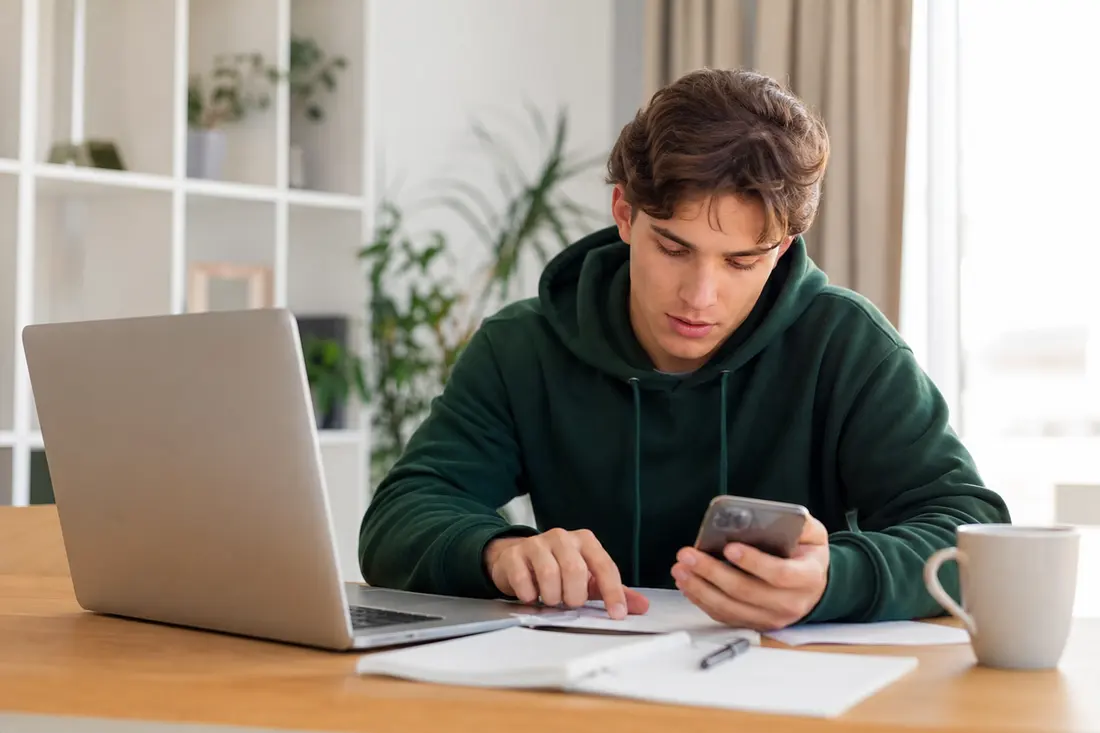 Jeune homme en sweat vert assis à un bureau lumineux, smartphone en main, qui pointe une feuille posée à côté de son ordinateur portable