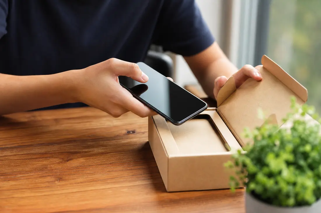 Un jeune homme sort un smartphone noir d'une boîte en carton kraft ouverte, posée sur une table en bois près d'une fenêtre