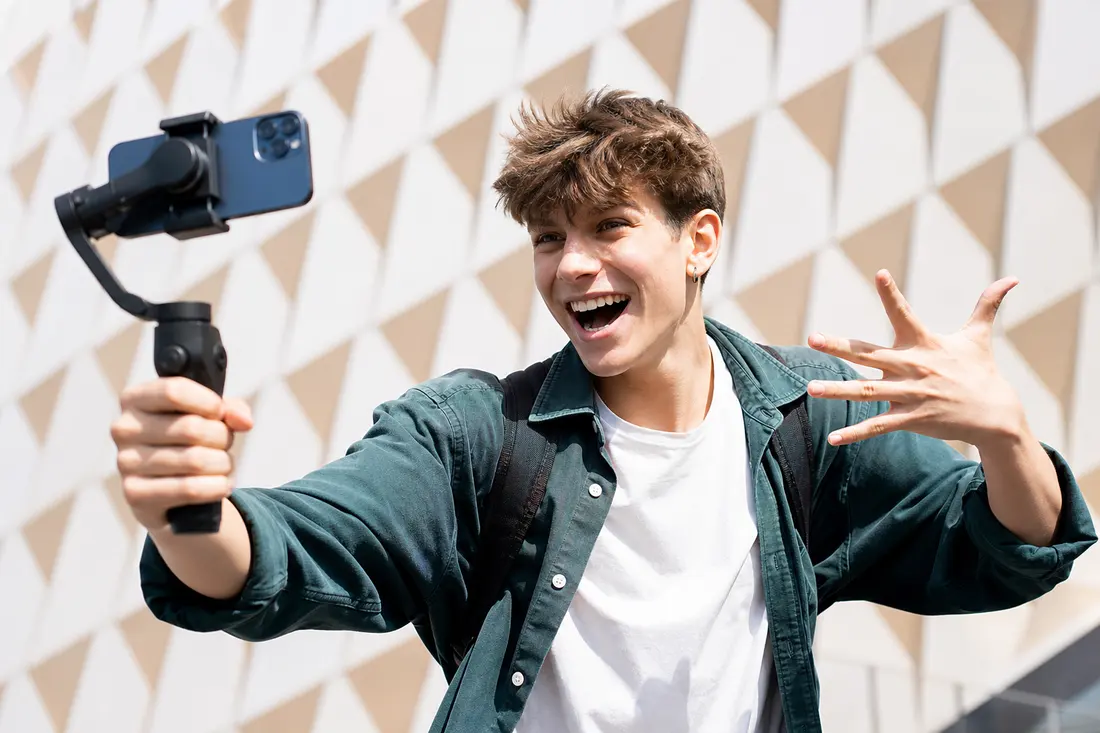 Jeune souriant filmant un selfie vidéo avec un smartphone sur stabilisateur, main levée, devant un mur géométrique en plein jour