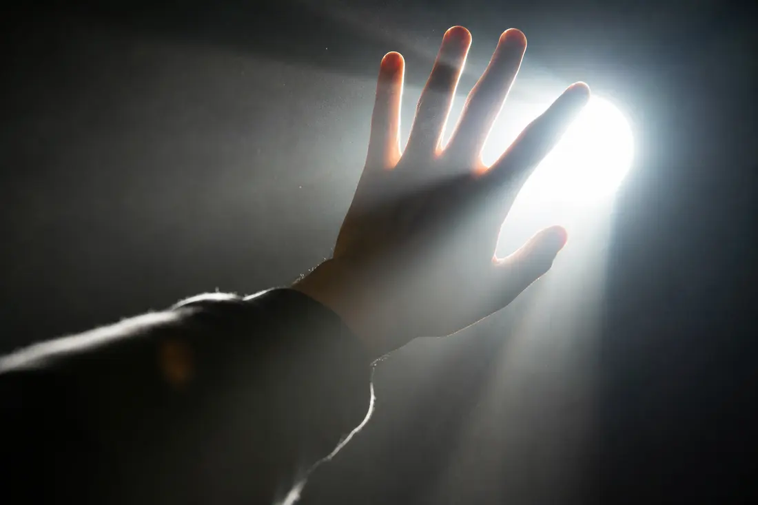 Une main tendue à contre-jour tente de masquer un faisceau lumineux intense, mais la lumière filtre entre les doigts dans une pièce sombre et poussiéreuse