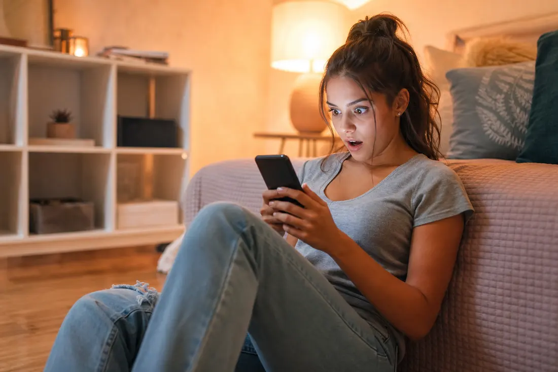 Jeune femme assise par terre contre son lit dans une chambre à la lumière chaude, smartphone dans les mains, bouche ouverte de surprise