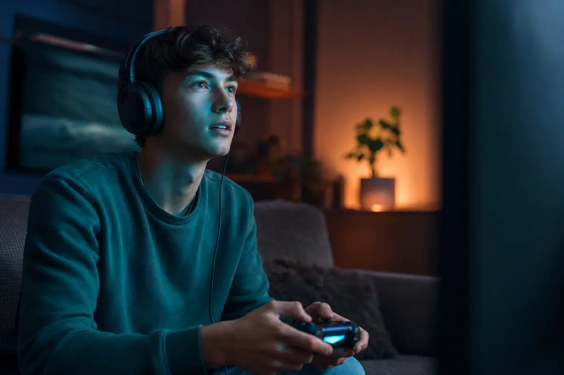 Jeune homme au casque audio, manette en main, assis sur un canapé dans un salon sombre éclairé par la lueur bleue de son écran