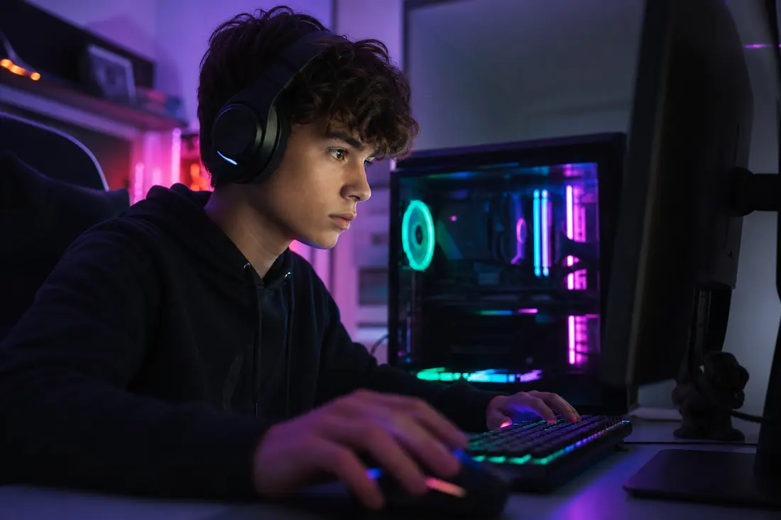 Un adolescent casque sur les oreilles, concentré devant un PC gamer aux LED violettes et vertes, les mains sur un clavier rétroéclairé