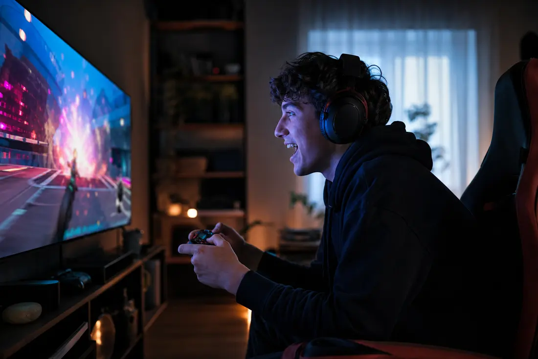 Jeune homme casque audio sur les oreilles et manette en main, souriant devant un grand écran de jeu dans un salon plongé dans la pénombre
