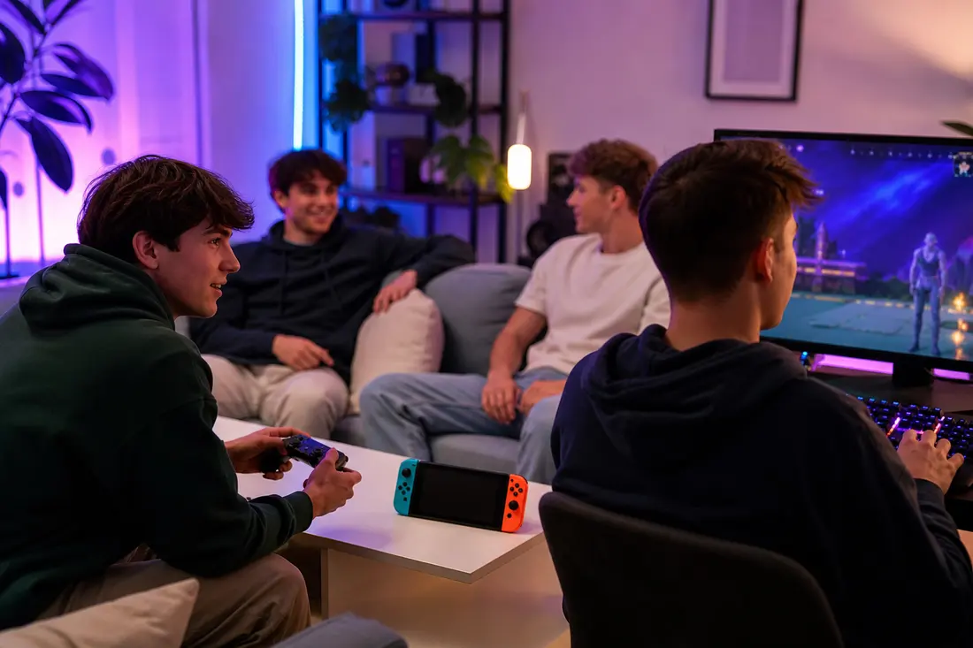 Quatre jeunes garçons en soirée jeu vidéo dans un salon aux lumières colorées, l'un tient une manette, un autre joue au clavier, console portable sur la table