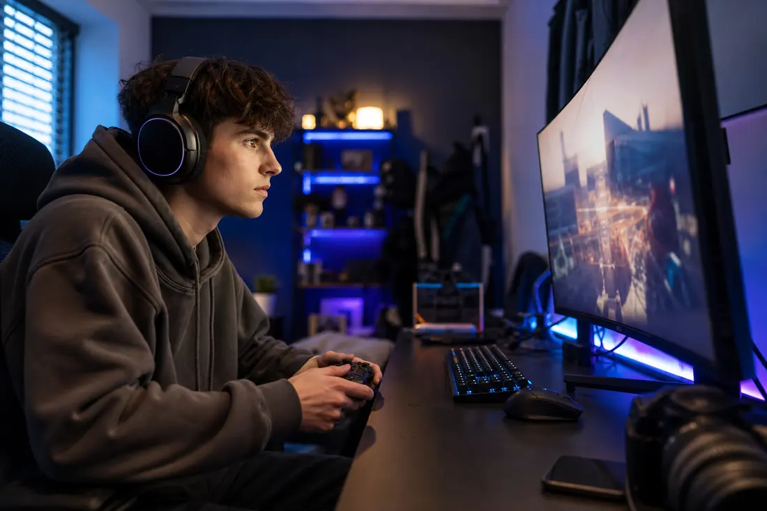 Jeune homme avec un casque audio et une manette en main, concentre devant un grand ecran incurve dans une chambre eclairee en bleu et violet
