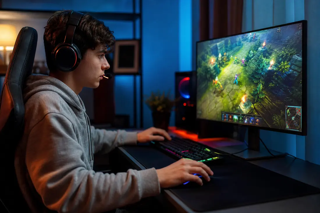 Un jeune joueur avec un casque-micro joue à un jeu multijoueur en ligne sur un PC de bureau, dans une chambre baignée de néons bleus et oranges