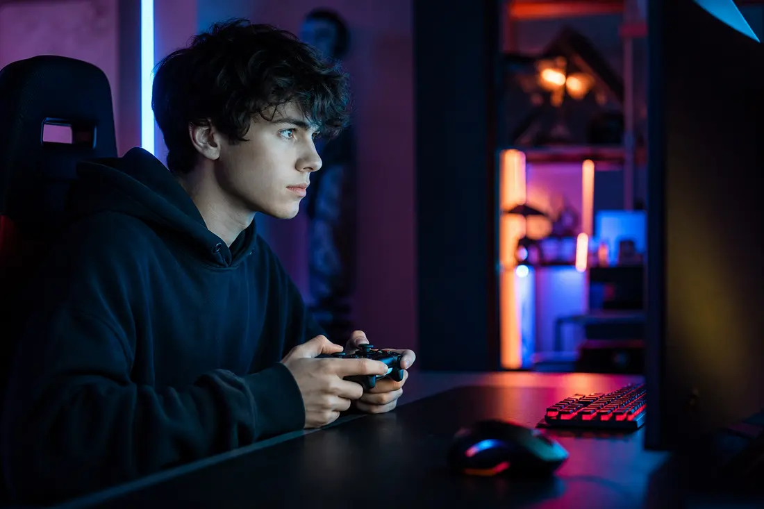 Un adolescent en sweat noir, manette en main, fixe son écran dans une chambre gaming éclairée de néons roses et bleus