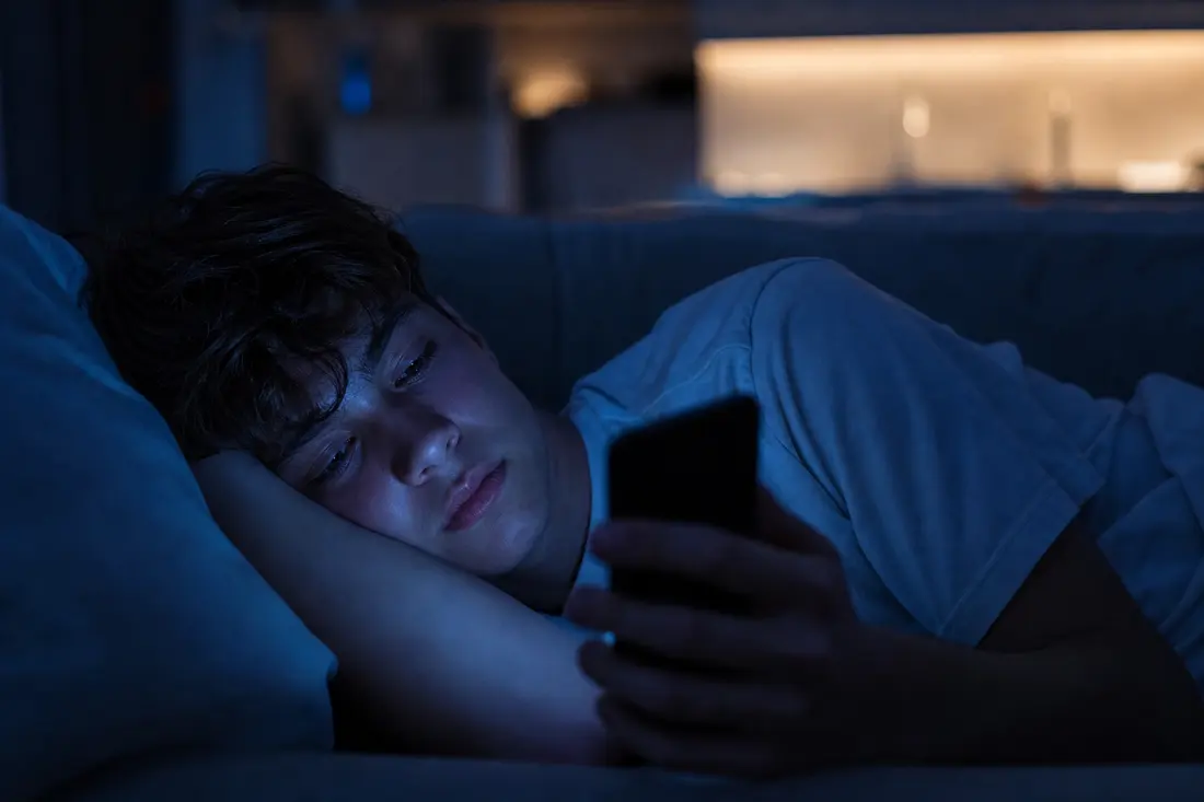 Adolescent allongé dans le noir sur un canapé, la tête posée sur son bras, le visage éclairé en bleu par l'écran du téléphone qu'il tient