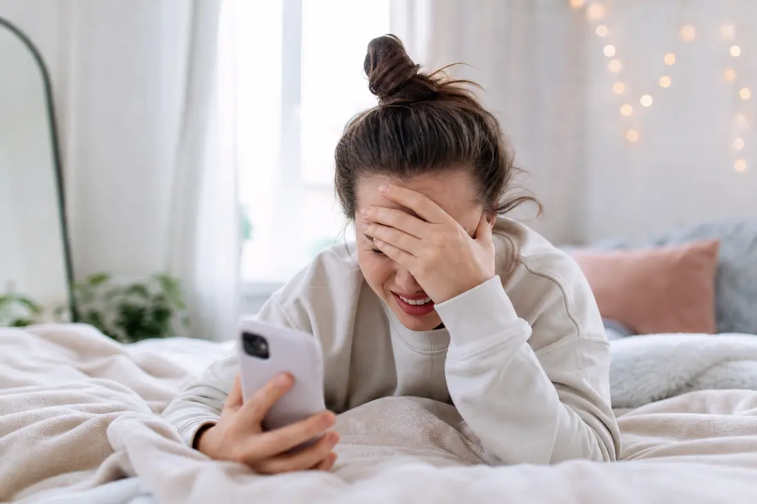 Jeune femme allongée sur un lit clair, une main sur les yeux et le sourire crispé, son téléphone dans l'autre main, dans une chambre lumineuse