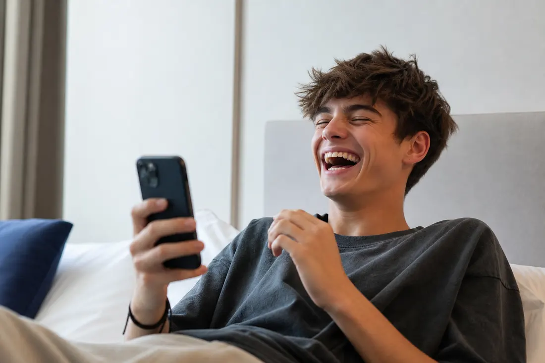 Adolescent allongé sur un lit, smartphone à la main, éclatant de rire les yeux fermés dans une chambre lumineuse