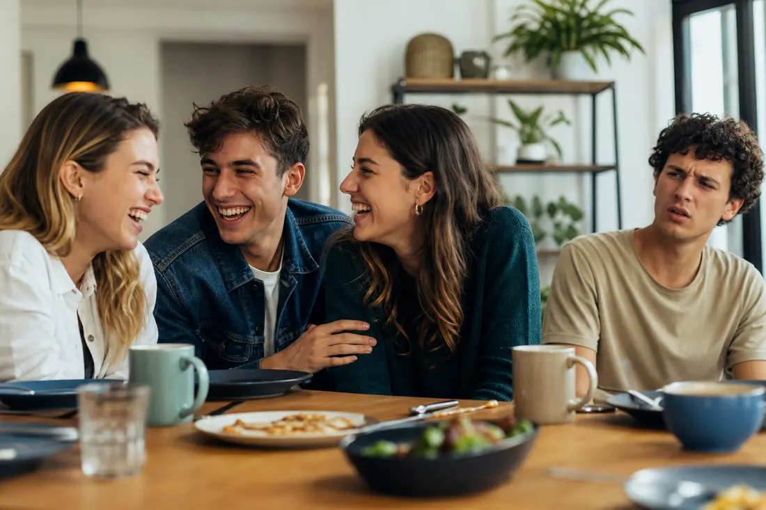Trois jeunes attablés autour d'un brunch rient aux éclats ensemble pendant qu'un quatrième, à droite, fronce les sourcils sans comprendre