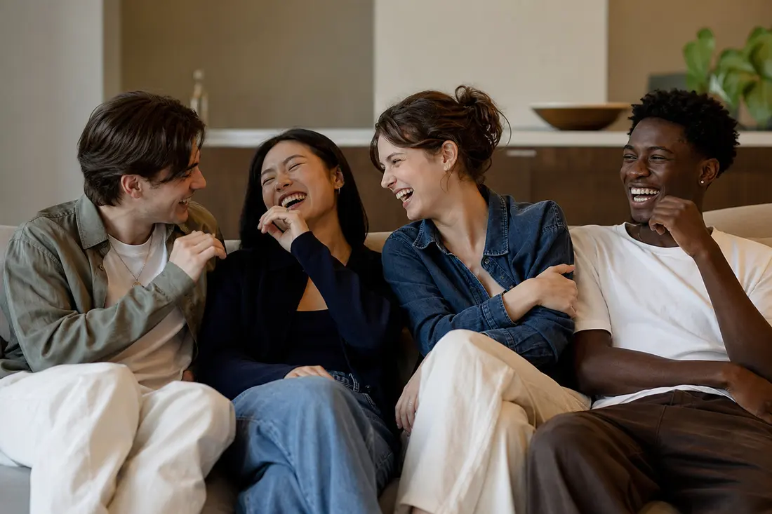 Quatre jeunes amis assis serrés sur un canapé dans un salon clair éclatent de rire ensemble en se regardant