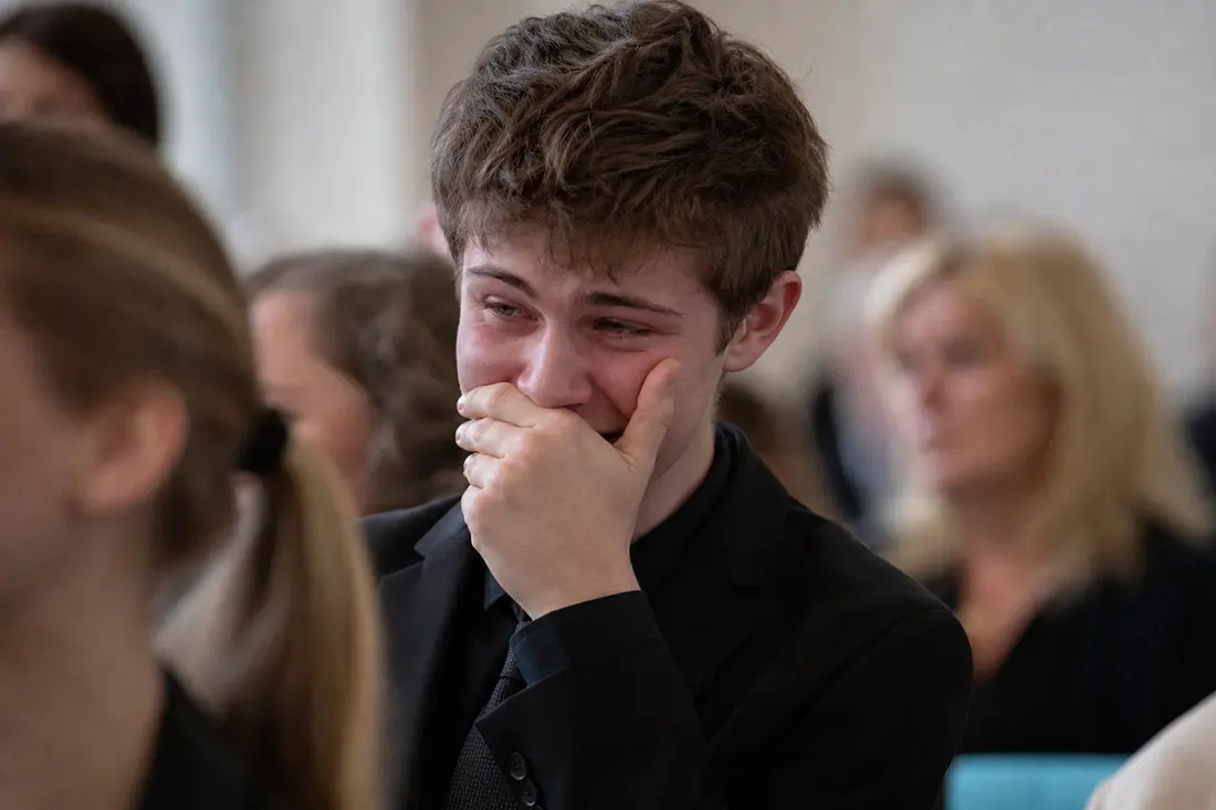 Jeune homme en costume noir assis lors d'une cérémonie, la main pressée sur la bouche et les yeux humides, entouré de personnes floues