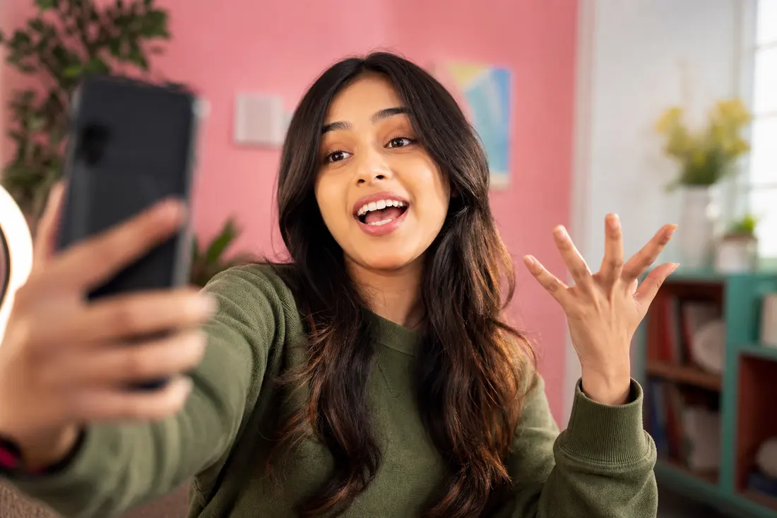 Jeune femme en sweat vert filmant une story en selfie avec son téléphone, bouche ouverte et main levée, devant un mur rose