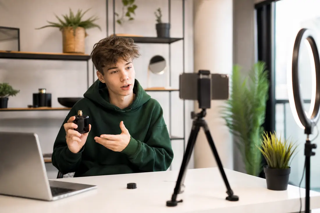 Jeune créateur en sweat vert filmant une vidéo avec son smartphone sur trépied, un petit flacon noir à la main, devant un anneau lumineux