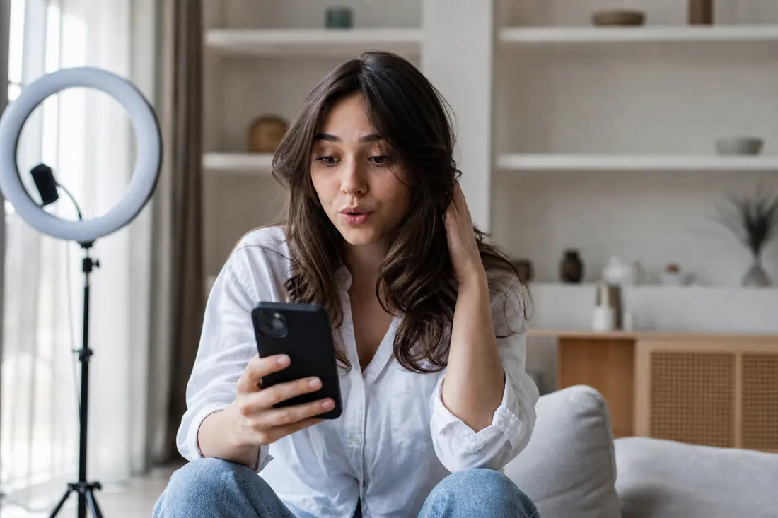Jeune femme brune assise dans un salon devant une ring light, l'air surpris, en train de regarder l'écran de son smartphone