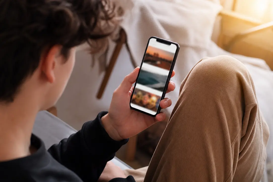 Un jeune vu de dos, assis dans un canapé, tient son smartphone et fait défiler un fil de photos dans une lumière chaude