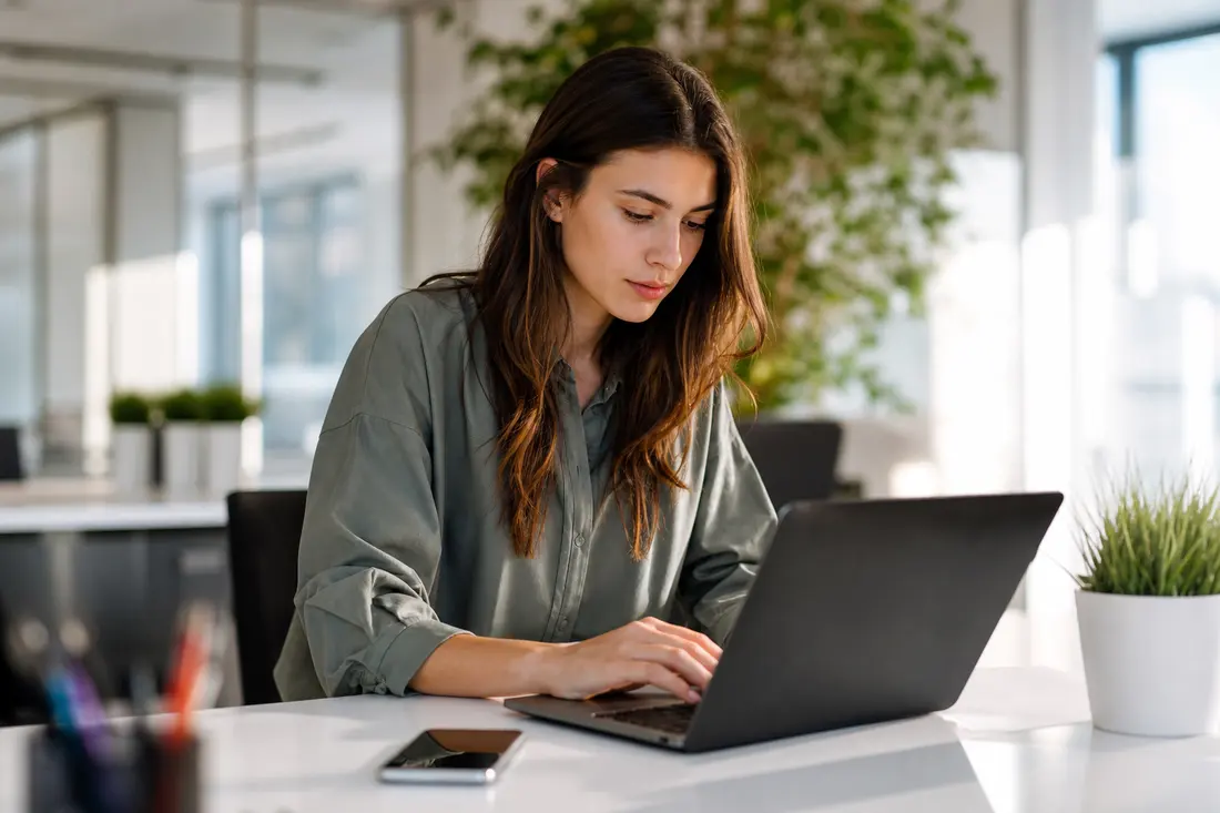 Jeune femme en chemise verte tapant sur un ordinateur portable dans un bureau lumineux, un smartphone posé à côté d'elle