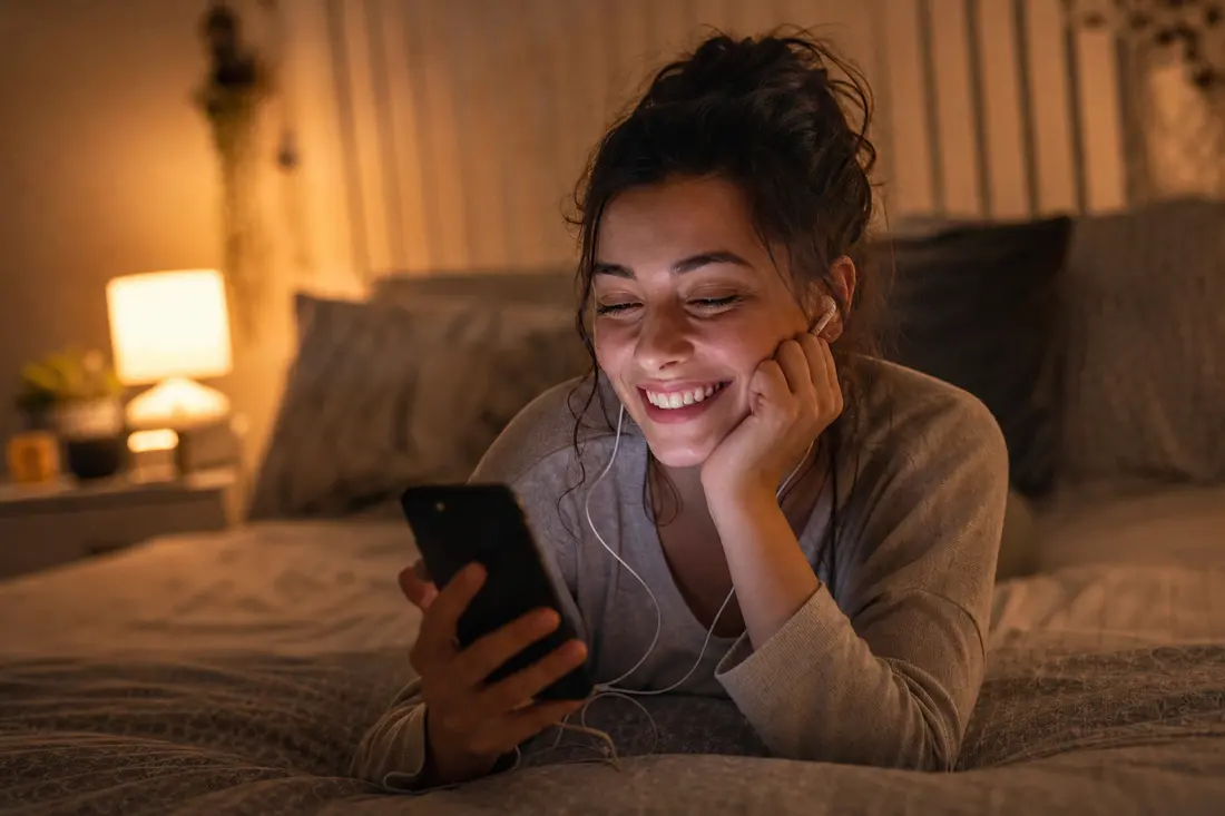 Jeune femme allongée sur son lit le soir, écouteurs aux oreilles, souriant devant son smartphone dans une chambre éclairée par une lampe chaude