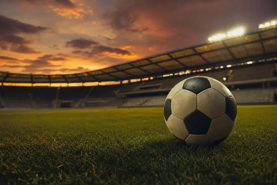 Ballon de football usé posé sur la pelouse d'un stade vide au coucher du soleil, tribunes et projecteurs allumés en arrière-plan
