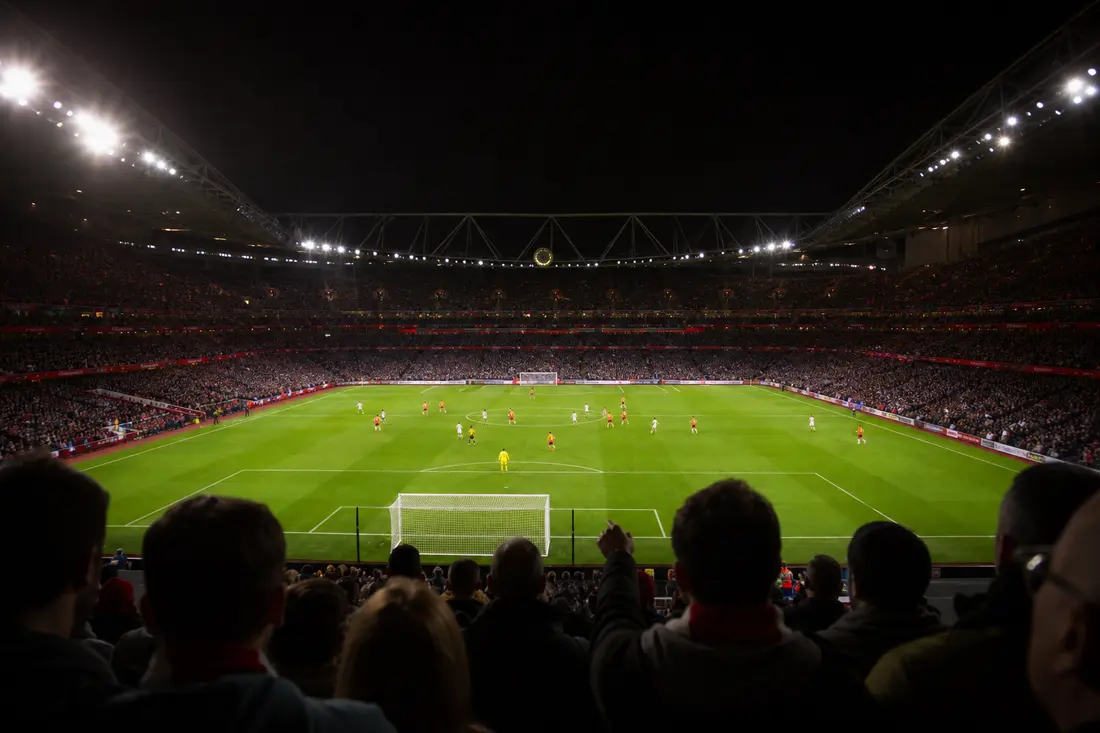 Vue depuis les tribunes d'un stade de football bondé en nocturne, pelouse éclairée par les projecteurs et joueurs en plein match
