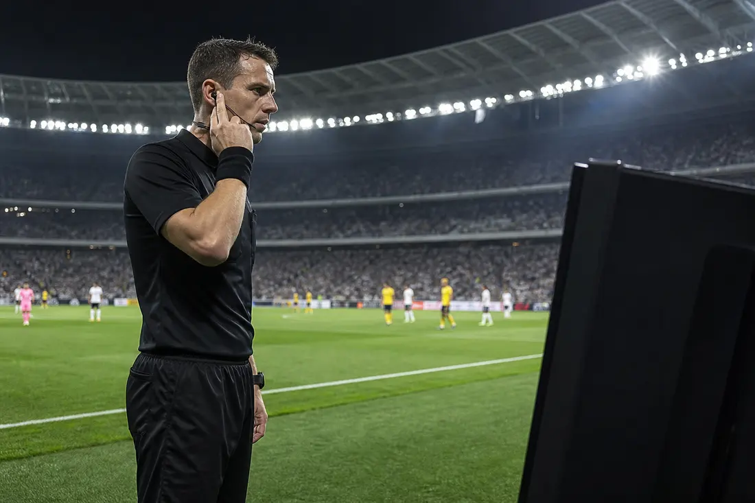 Un arbitre de football en tenue noire, la main sur son oreillette, se tient devant l'écran de la VAR au bord d'une pelouse, dans un stade plein la nuit