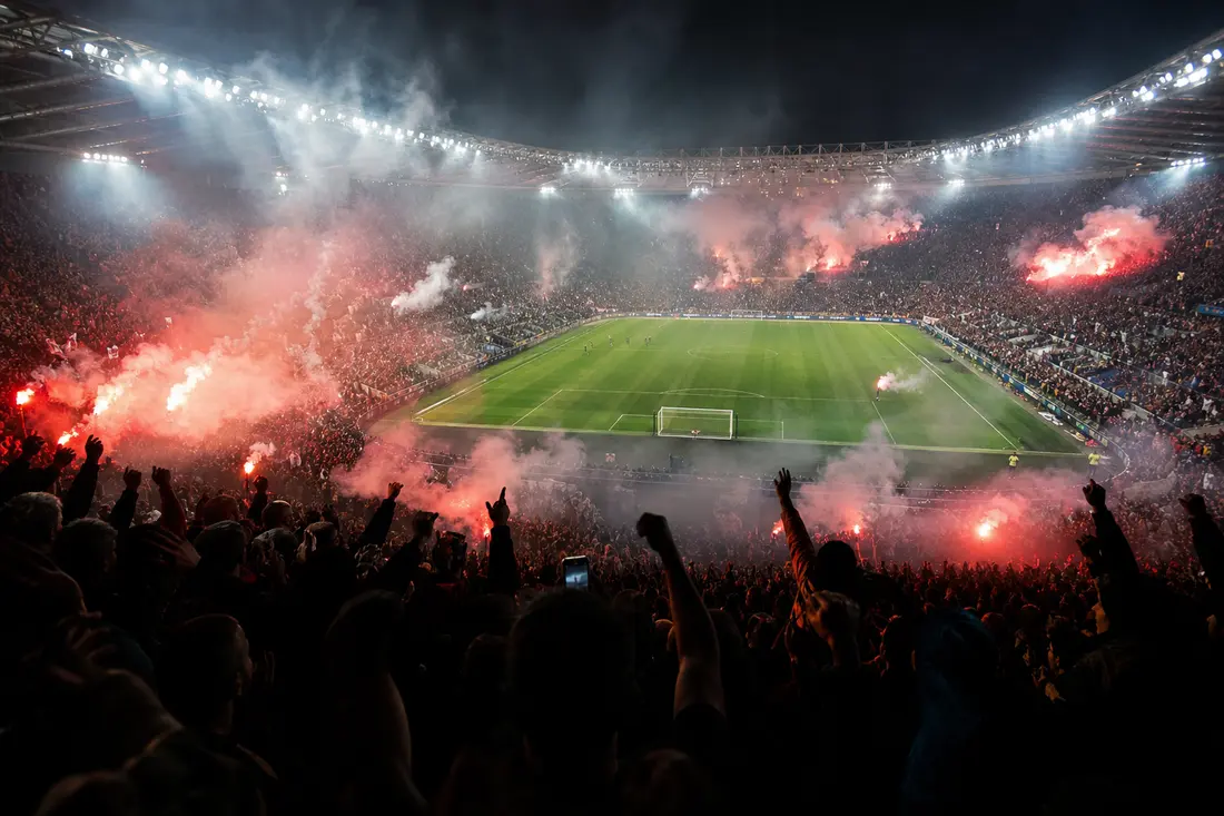 Stade de football bondé la nuit, tribunes noyées dans la fumée rouge des fumigènes et bras de supporters levés vers la pelouse éclairée