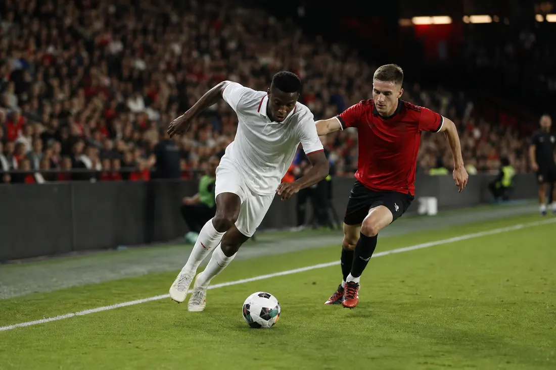 Deux footballeurs en duel de vitesse le long de la ligne de touche, maillot blanc contre maillot rouge, ballon au sol sous les projecteurs du stade
