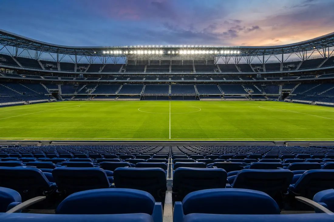 Stade de football désert au crépuscule, pelouse verte éclairée par les projecteurs et rangées de sièges bleus totalement vides au premier plan