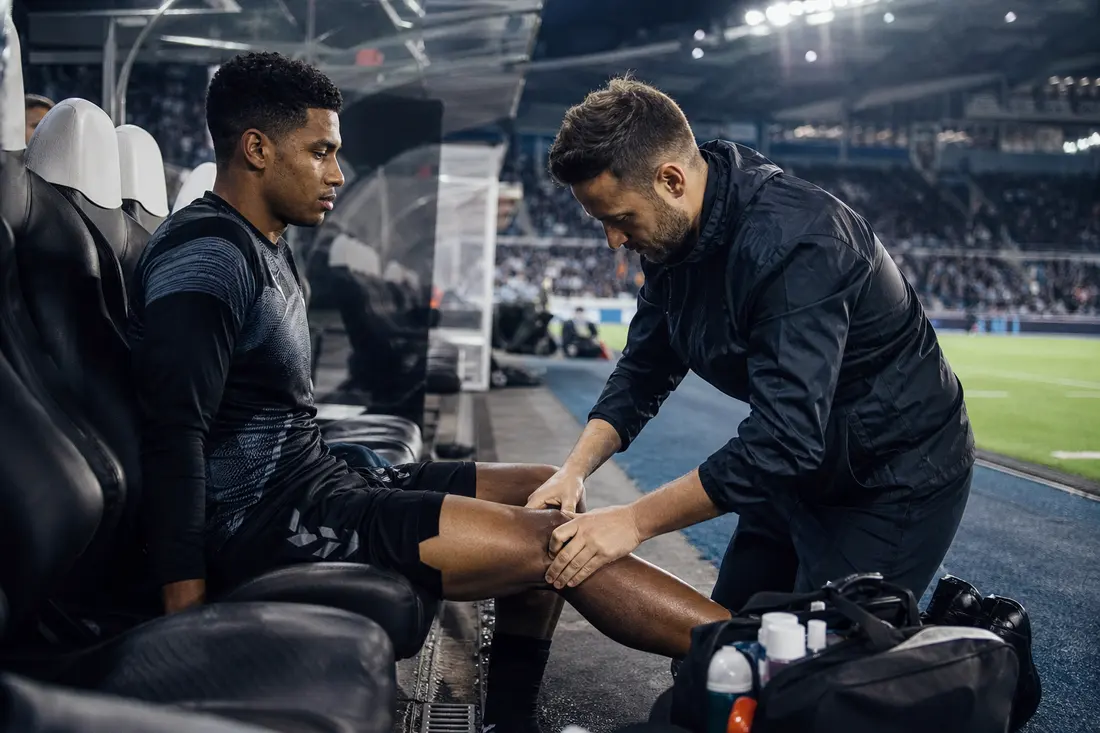 Un jeune footballeur assis sur le banc de touche pendant qu'un membre du staff médical examine son genou, stade éclairé en arrière-plan