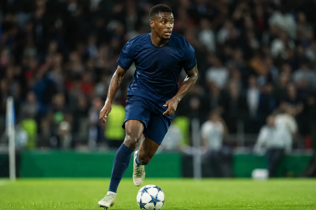 Jeune footballeur en maillot bleu marine, ballon au pied, en pleine course sur la pelouse d'un stade éclairé, public flou derrière lui