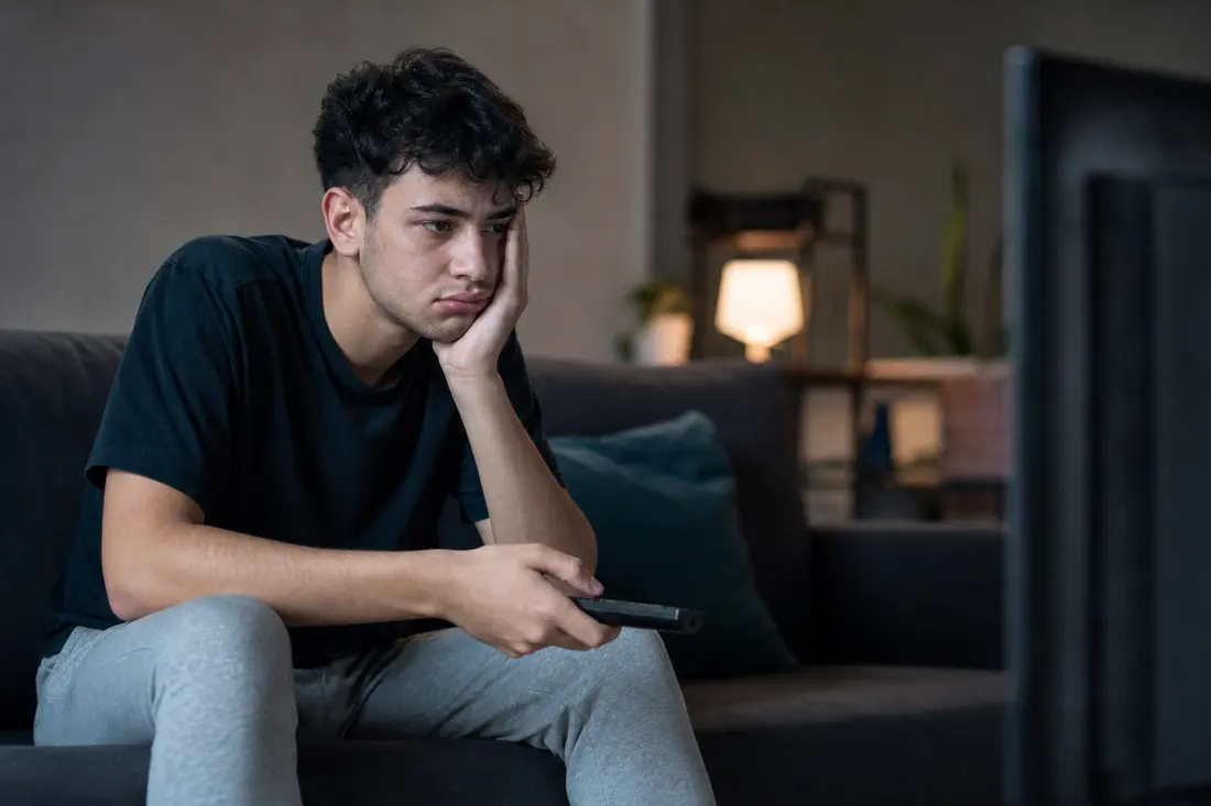 Jeune homme à l'air abattu, assis sur un canapé dans un salon tamisé, la télécommande à la main, le regard fixé sur un écran de télévision
