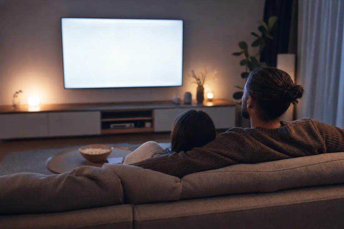 Un couple vu de dos sur un canapé dans un salon sombre, face à un grand écran de télévision allumé, avec un bol de popcorn sur la table basse