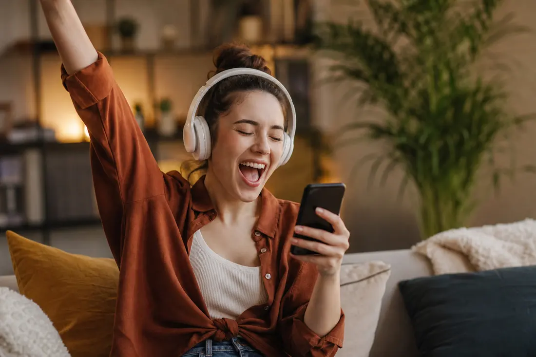 Jeune femme au casque audio blanc assise sur un canapé, bras levé et bouche ouverte de joie, son smartphone à la main dans un salon chaleureux
