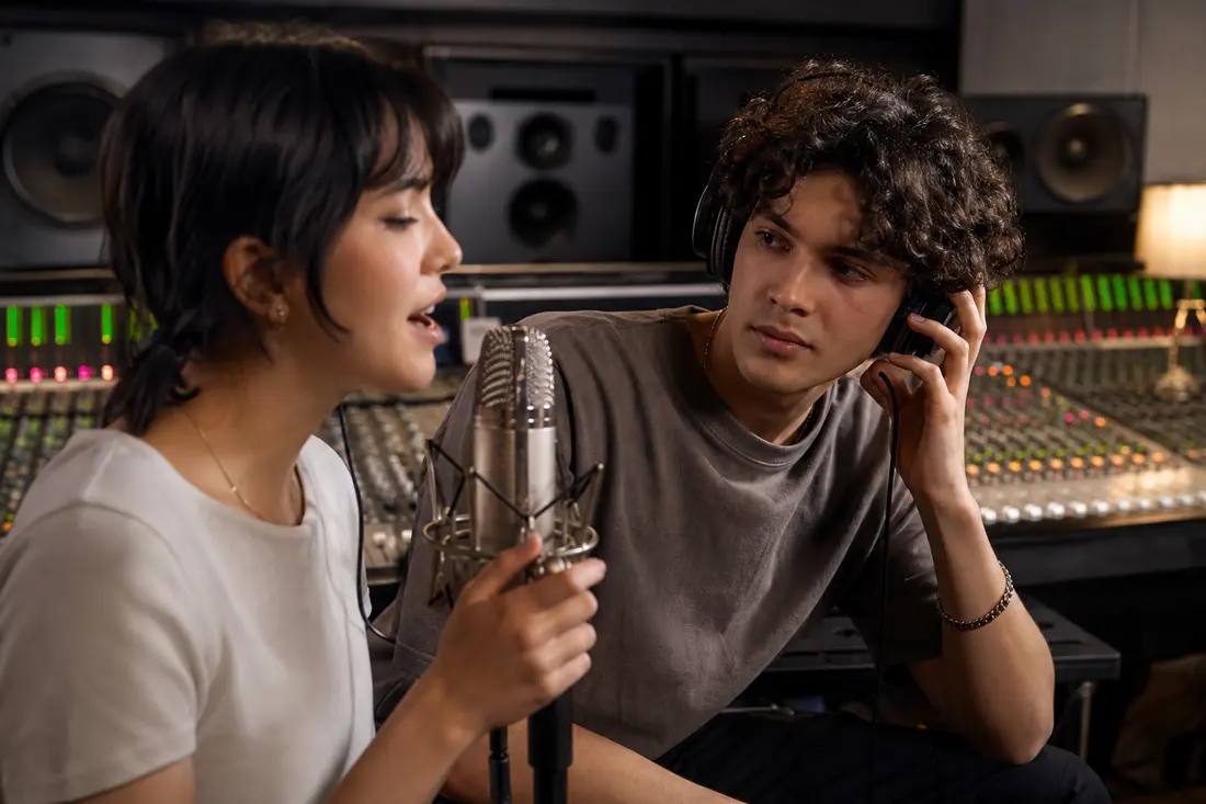 Une jeune femme chante devant un micro de studio pendant qu'un jeune homme, casque sur une oreille, l'écoute devant la console de mixage