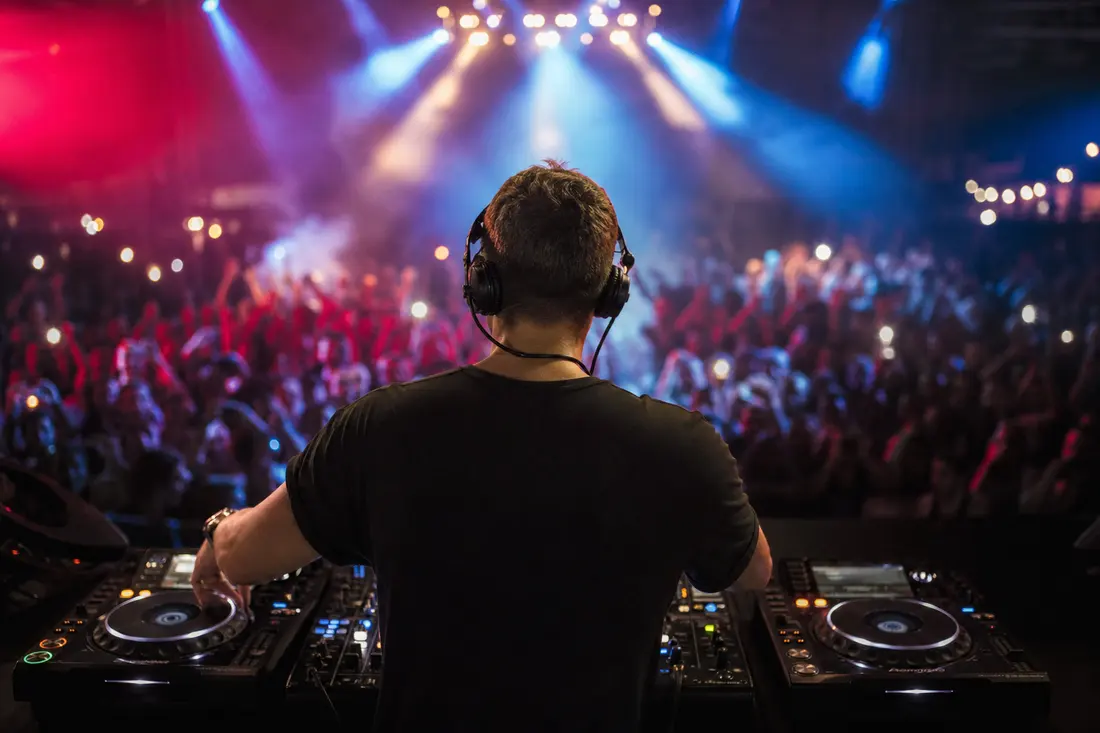 DJ de dos, casque sur les oreilles, mains sur deux platines face à une foule aux bras levés sous des lumières rouges et bleues