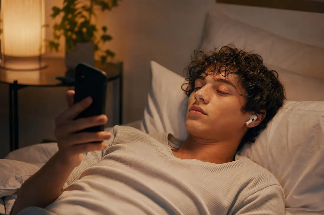 Jeune homme aux cheveux bouclés allongé dans son lit le soir, un écouteur sans fil à l'oreille, le regard fixé sur l'écran de son téléphone