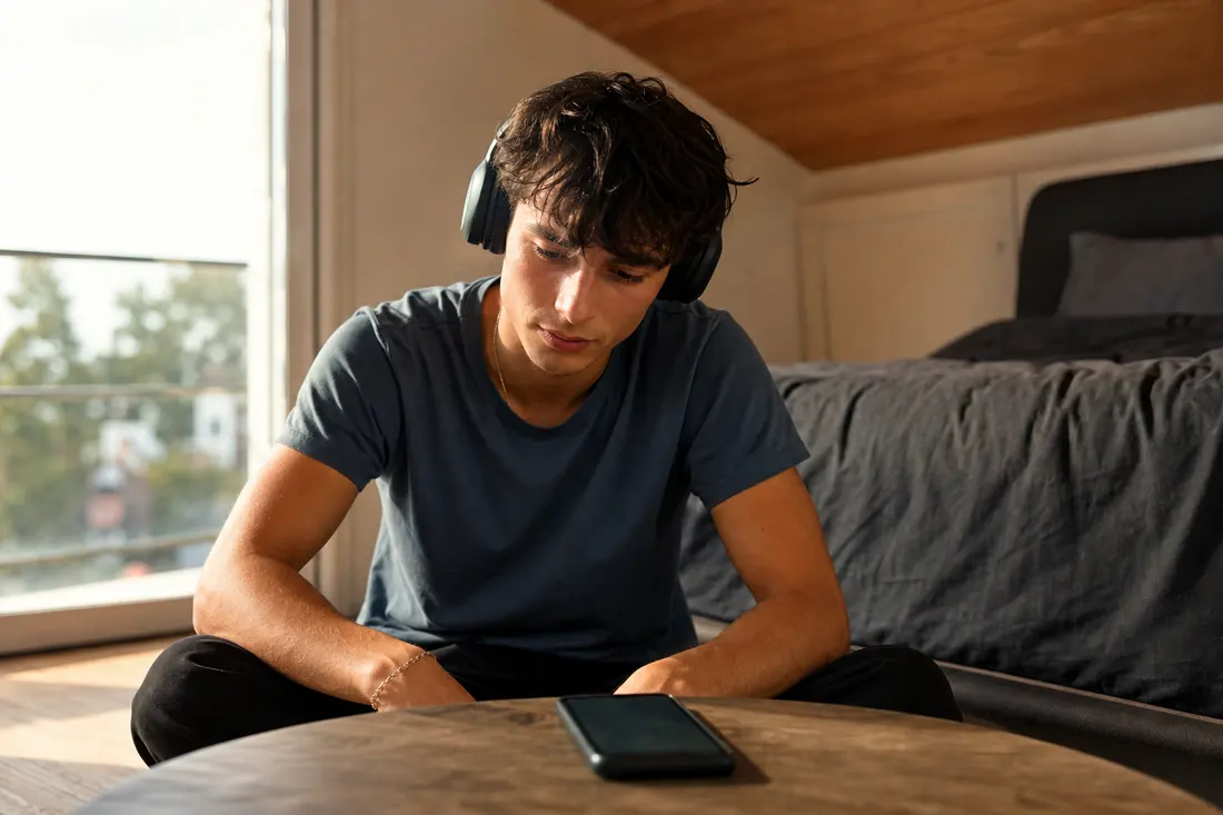 Jeune homme au casque audio assis en tailleur dans une chambre lumineuse, penché vers son smartphone posé sur une table basse ronde en bois